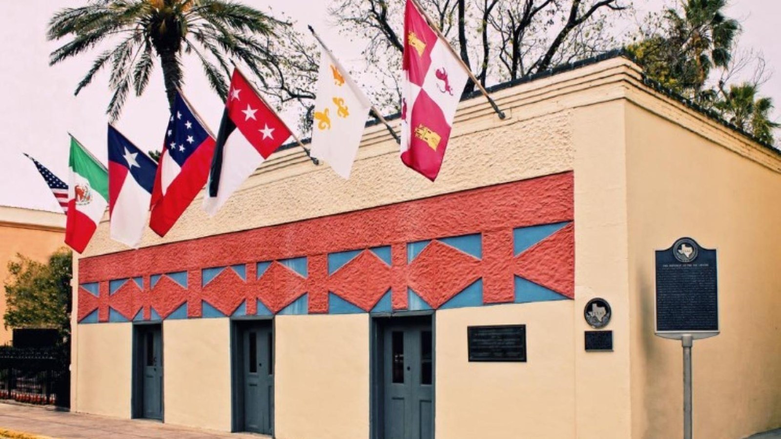 A tan building with three doors and a red and blue design above them. Seven flags line the top 