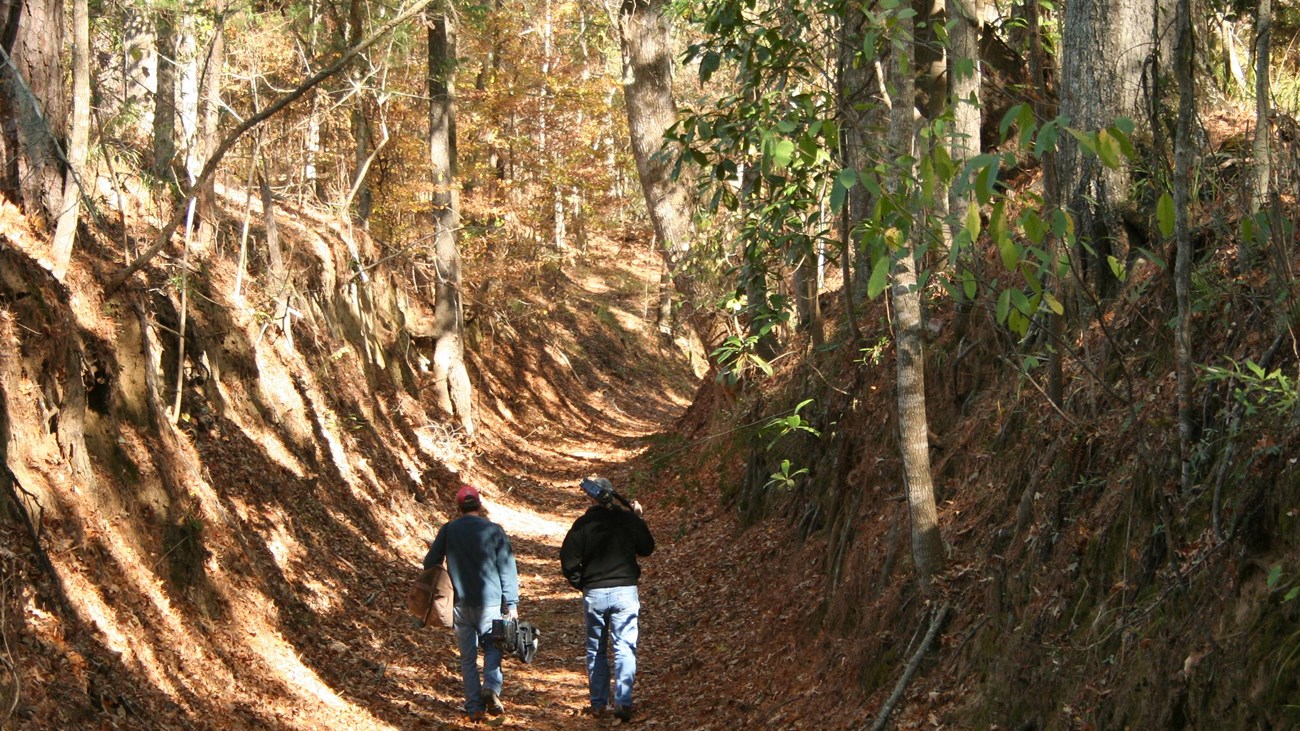 Two hikers walking away from viewer. Trail in gully with 20 foot embankments on each side.