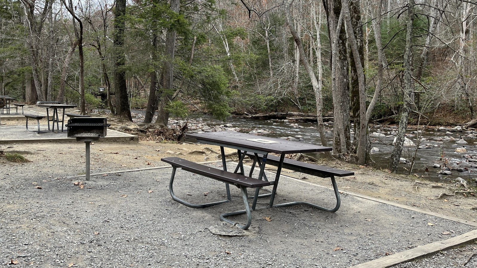 A brown picnic table with metal legs beside a metal grill on a gravel pad along a creek in winter.