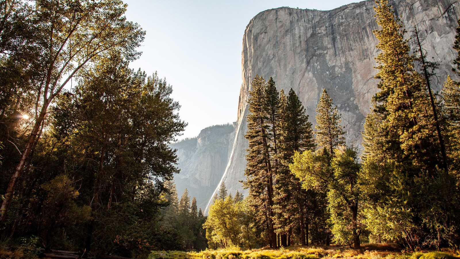 A giant granite monolith looms over trees, a river, and a sandy beach