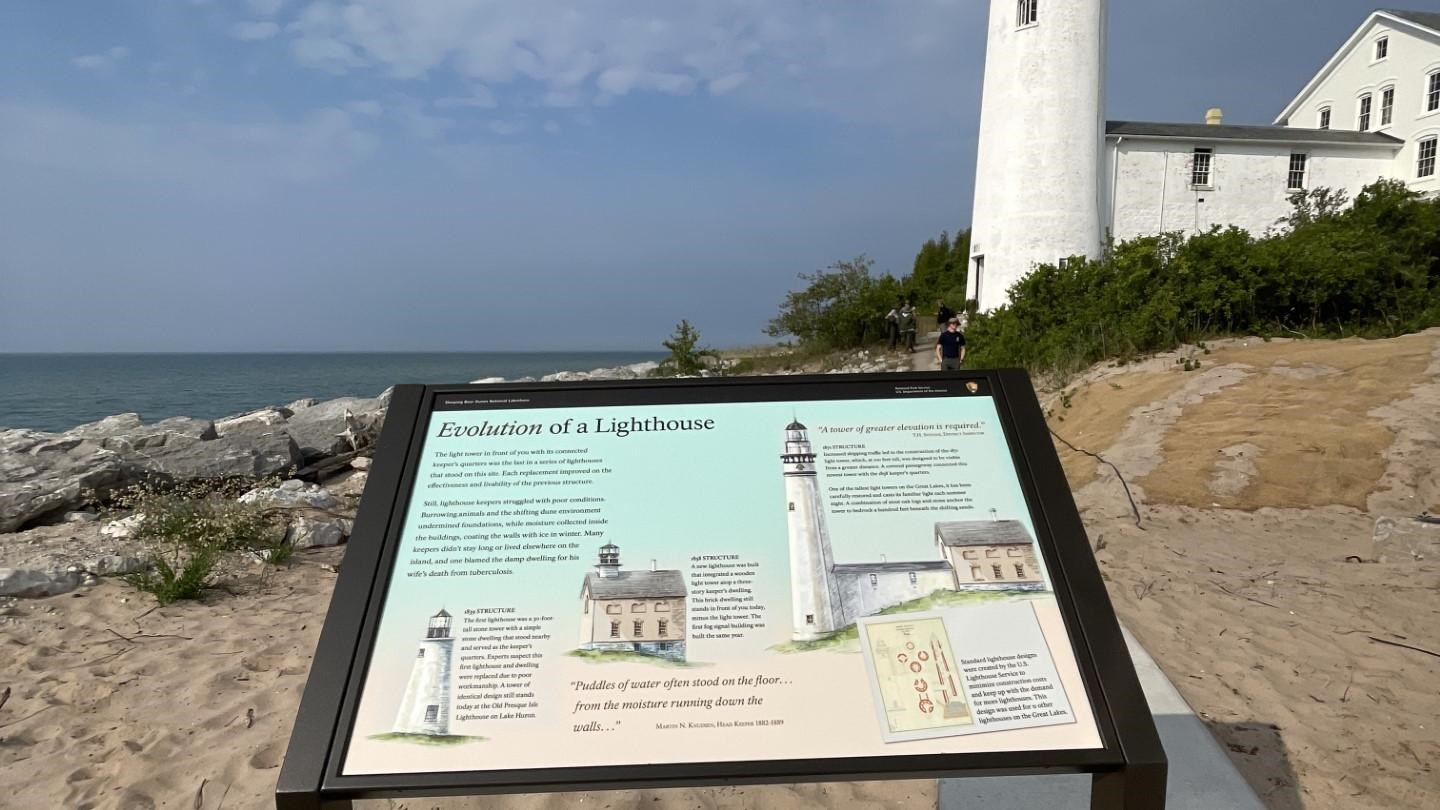 A cantilever wayside on a concrete platform. A white lighthouse and historic building are behind