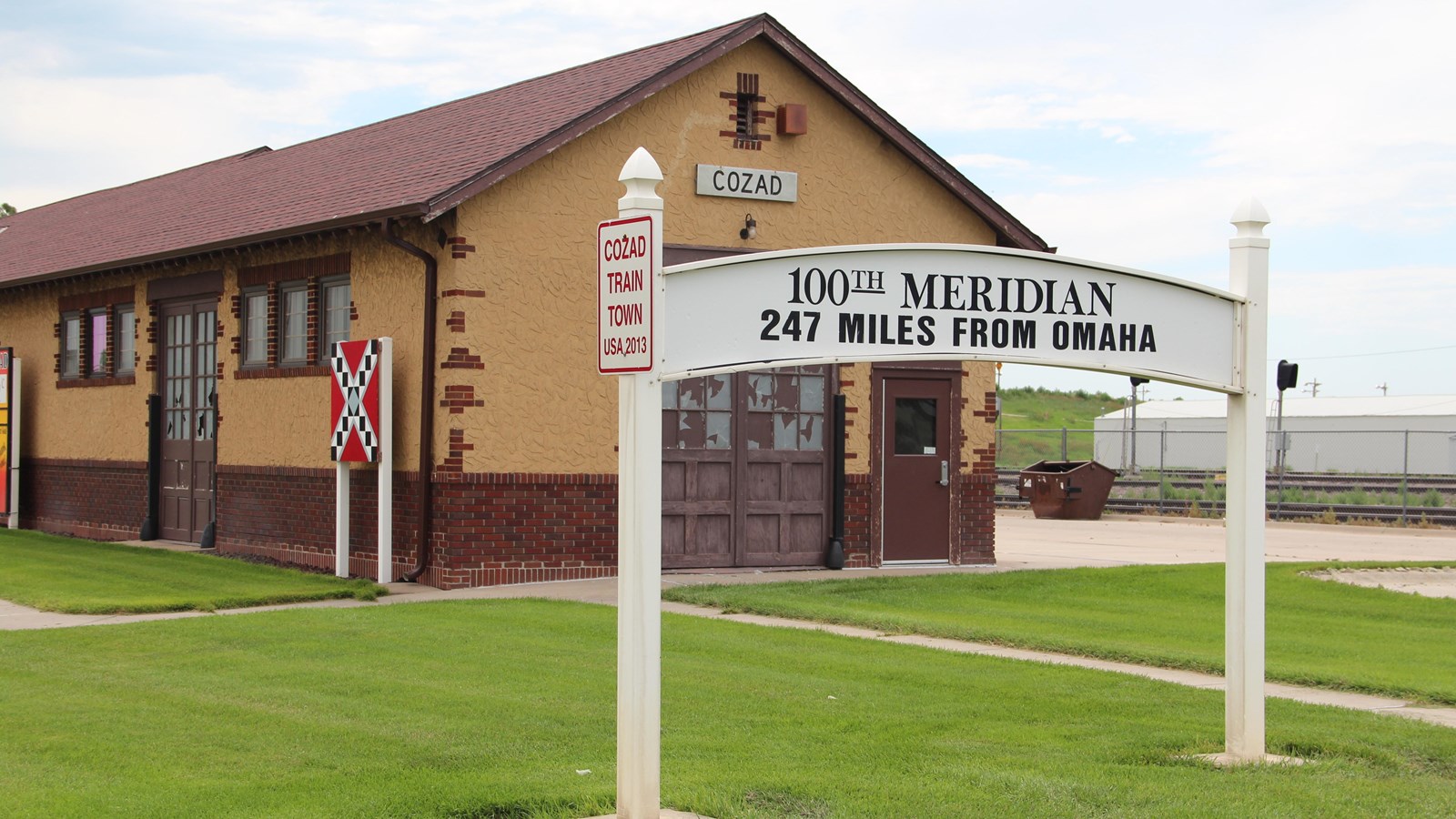  On a lush, green lawn in the foreground stands an arched, white sign between 2 wooden posts.
