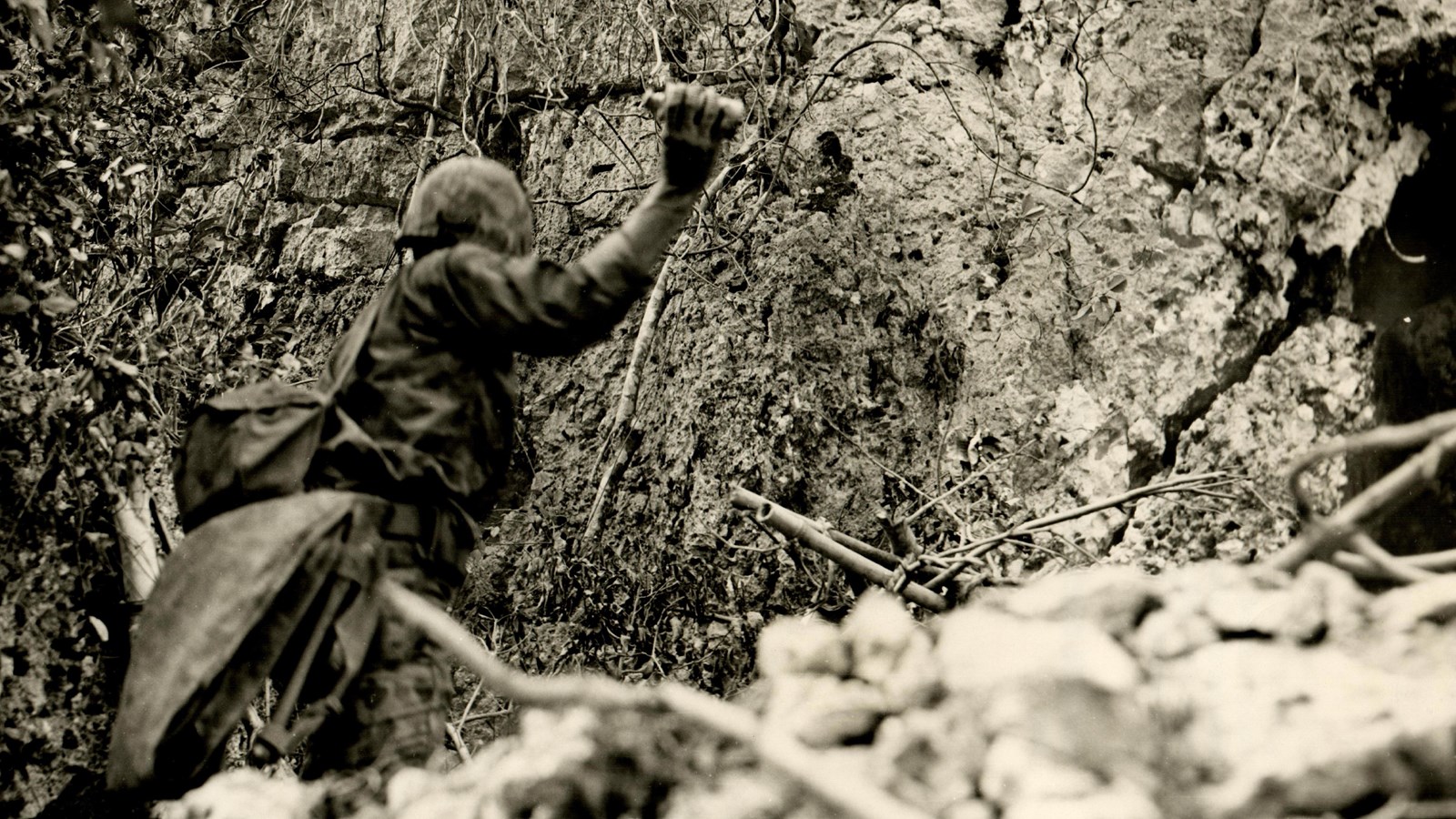 A marine has his arm back preparing to throw a grenade in his right hand. He\'s facing a cliffside.