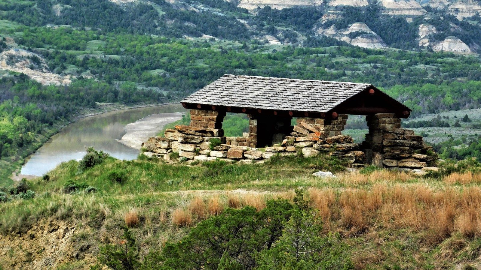 A small stone shelter with a low pitched roof stands at the edge of a butte overlooking a river.