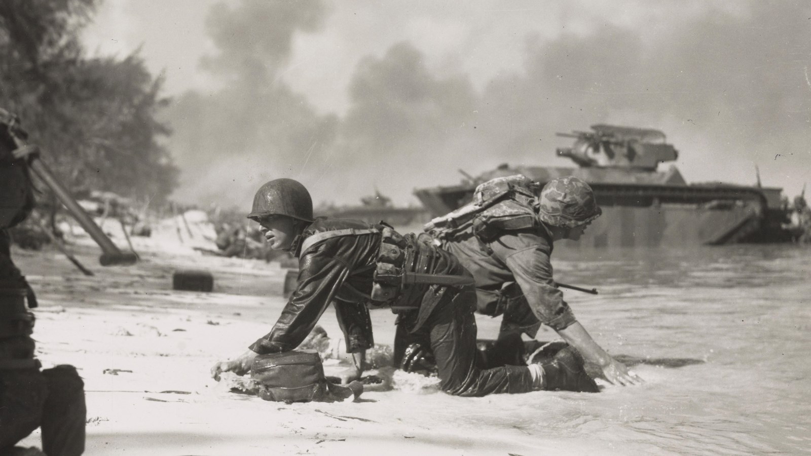 Two Marines crawling on the beach. They have helmets on, supply packs and weapons strapped to them.