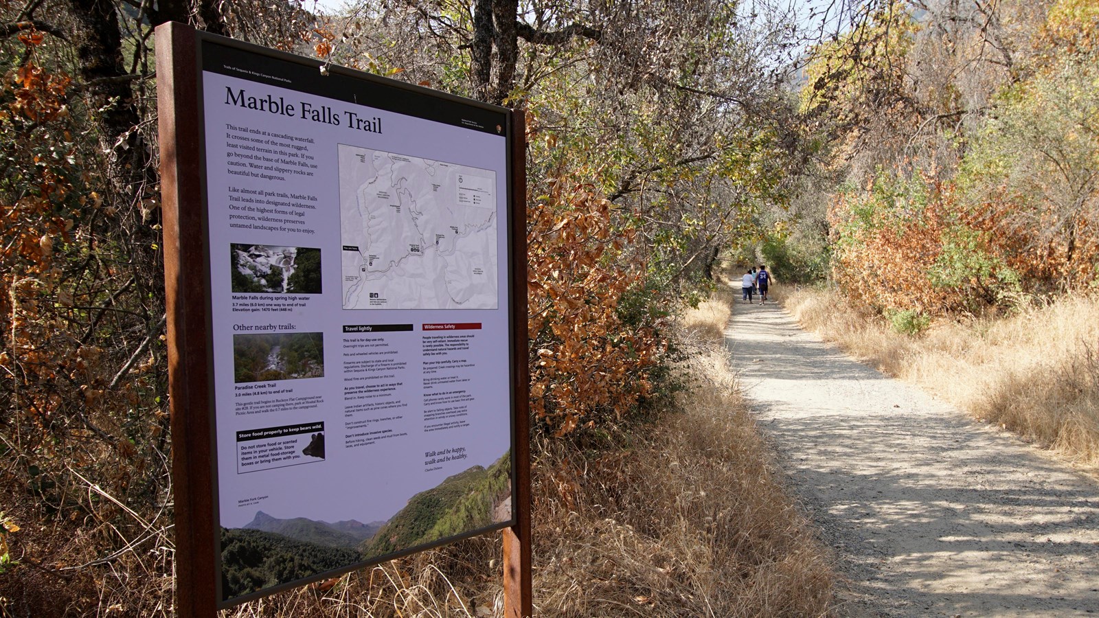 A blue metal panel sits in a metal frame next to a trail behind it are yellow grass and oak tree