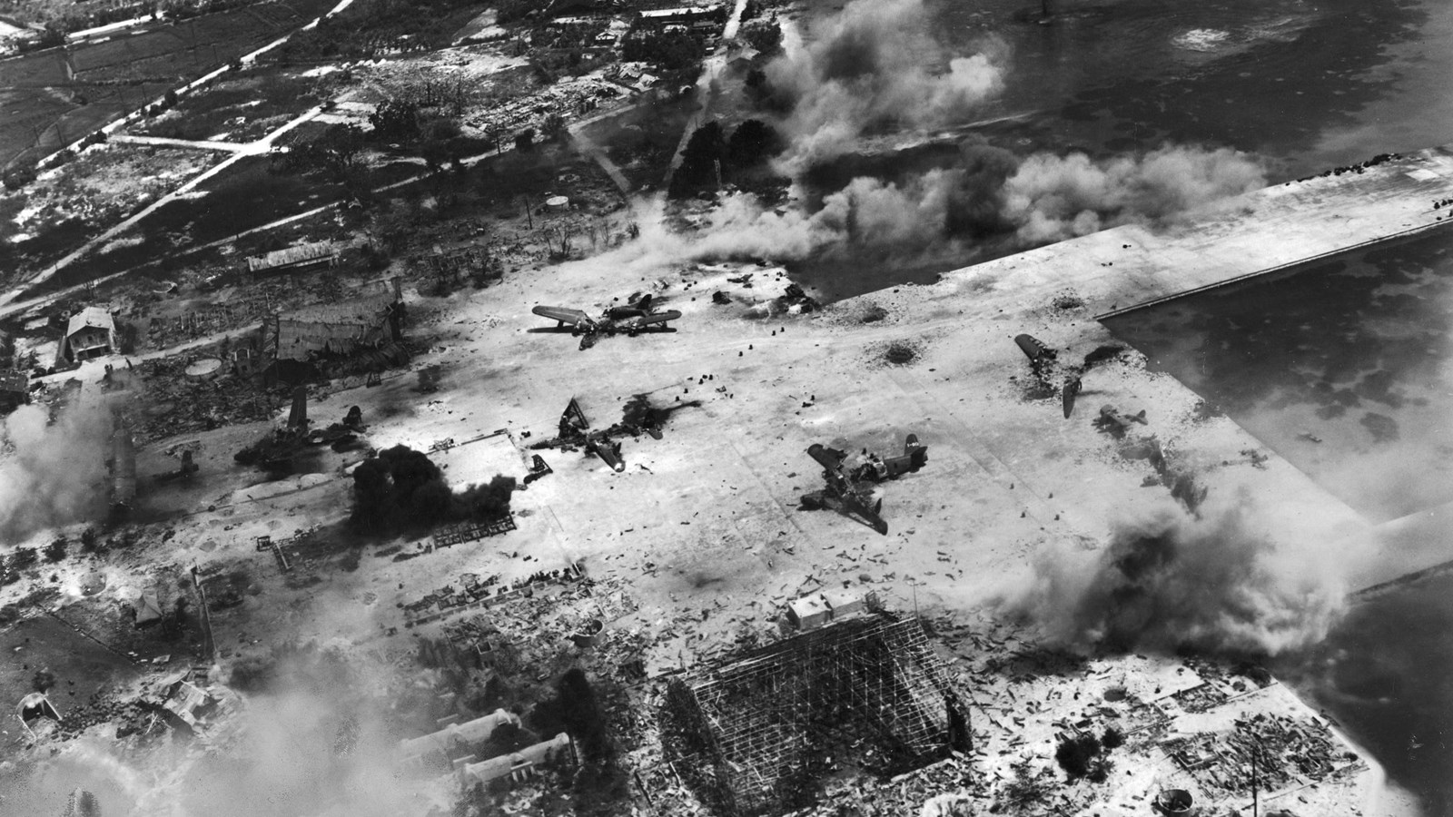 War ravaged airfield with billowing smoke from bombs. Ships in the ocean are in the distance.
