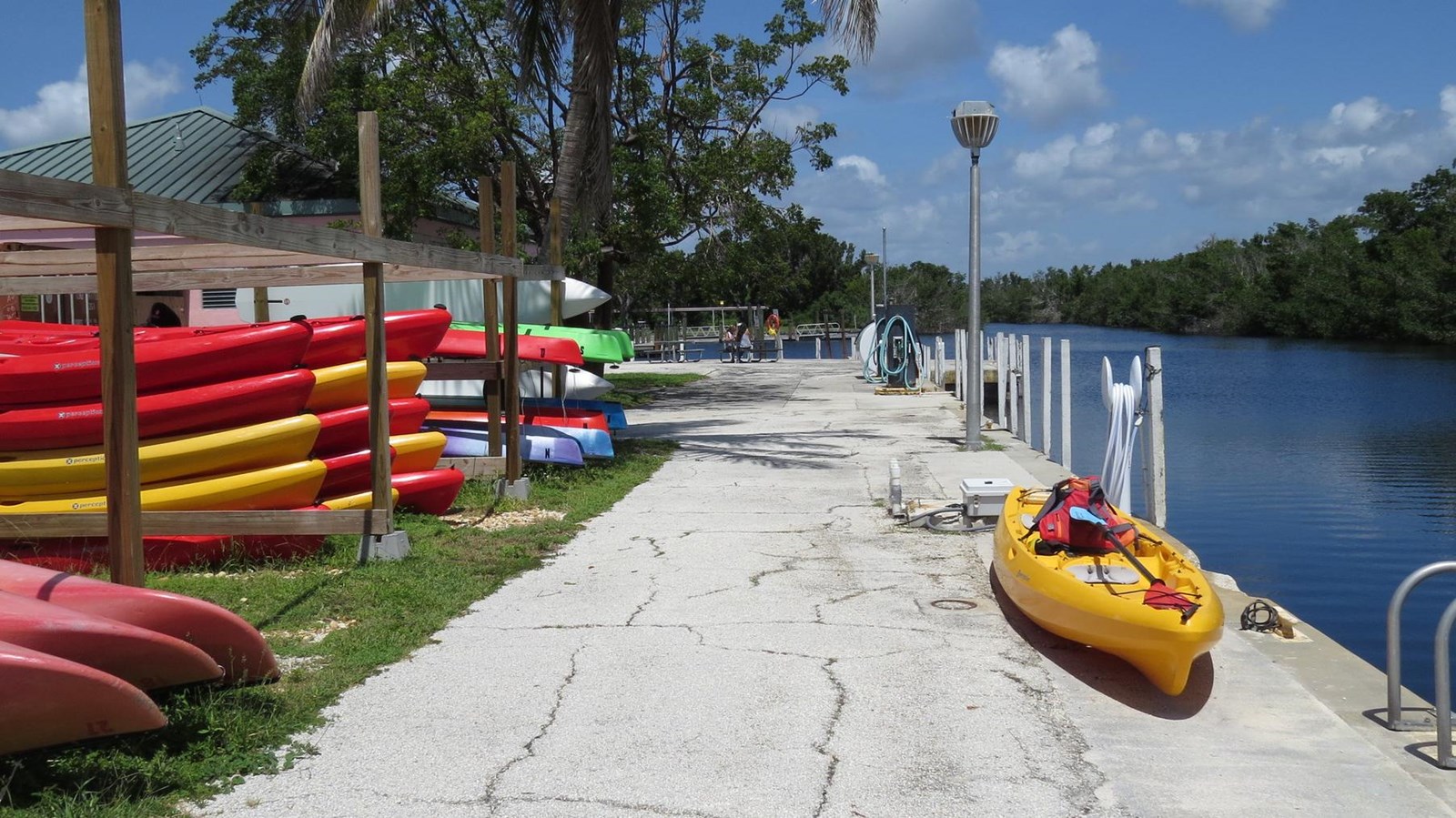 Colorful red and yellow kayaks are stacked along the grass next to a cement wall and blue water