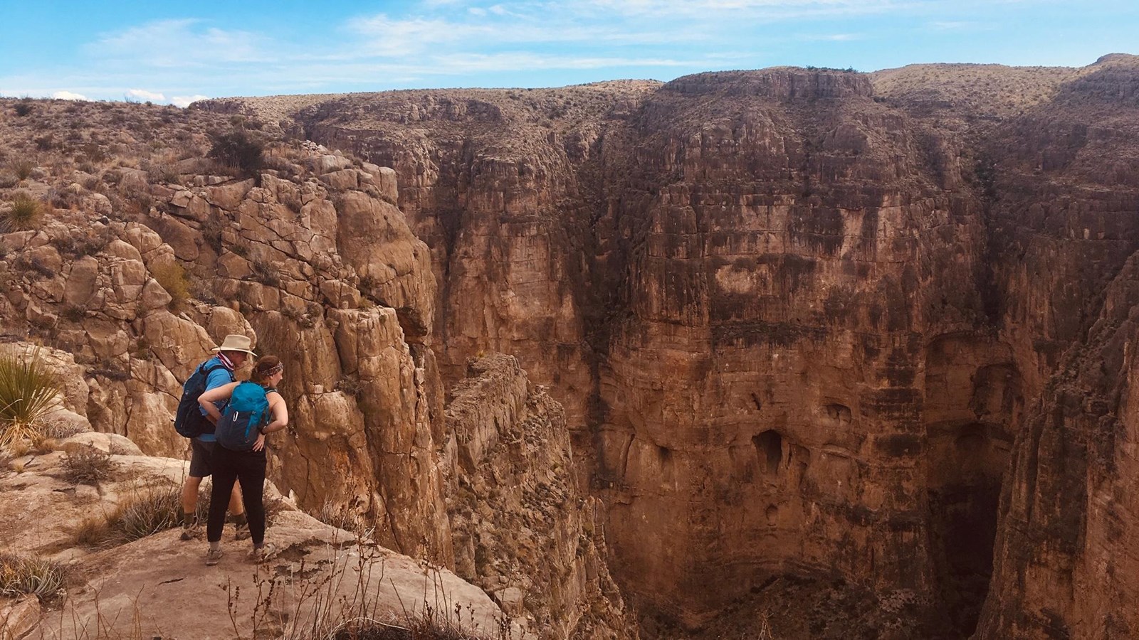 Two hikers peer over the edge of a high limestone cliff.