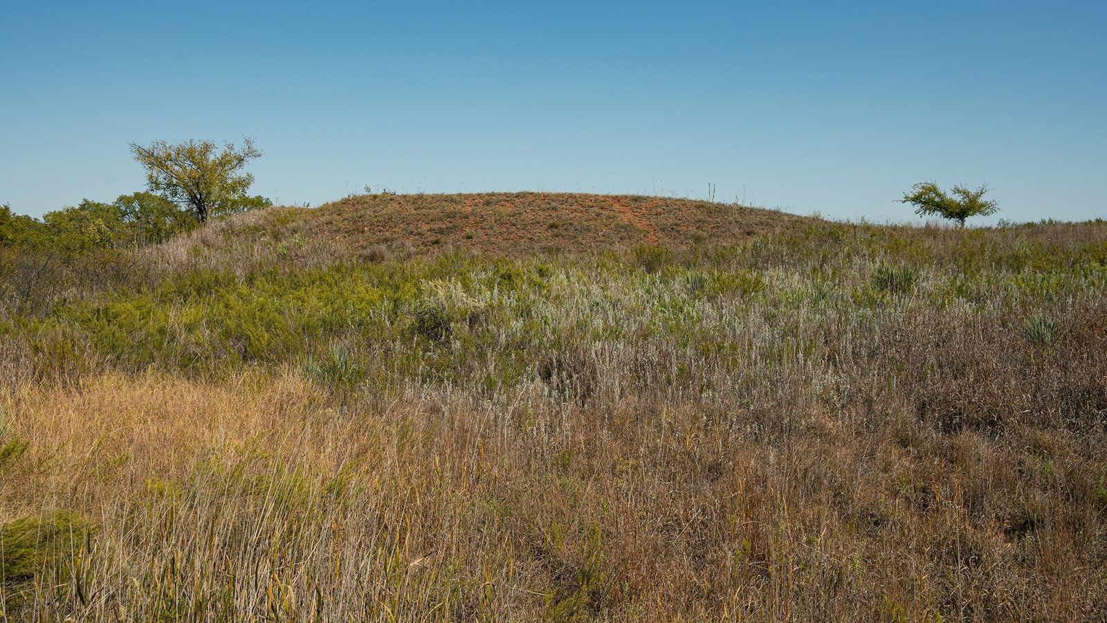 A small red hill rising out of a grassy area under a blue sky.