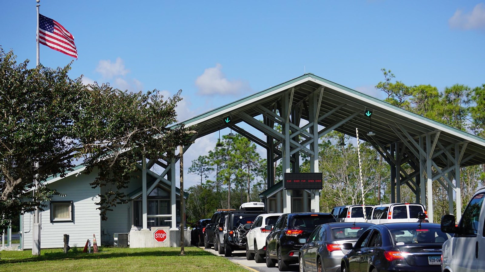 A line of cars waiting to enter through a gray open aired structure with a slanted roof