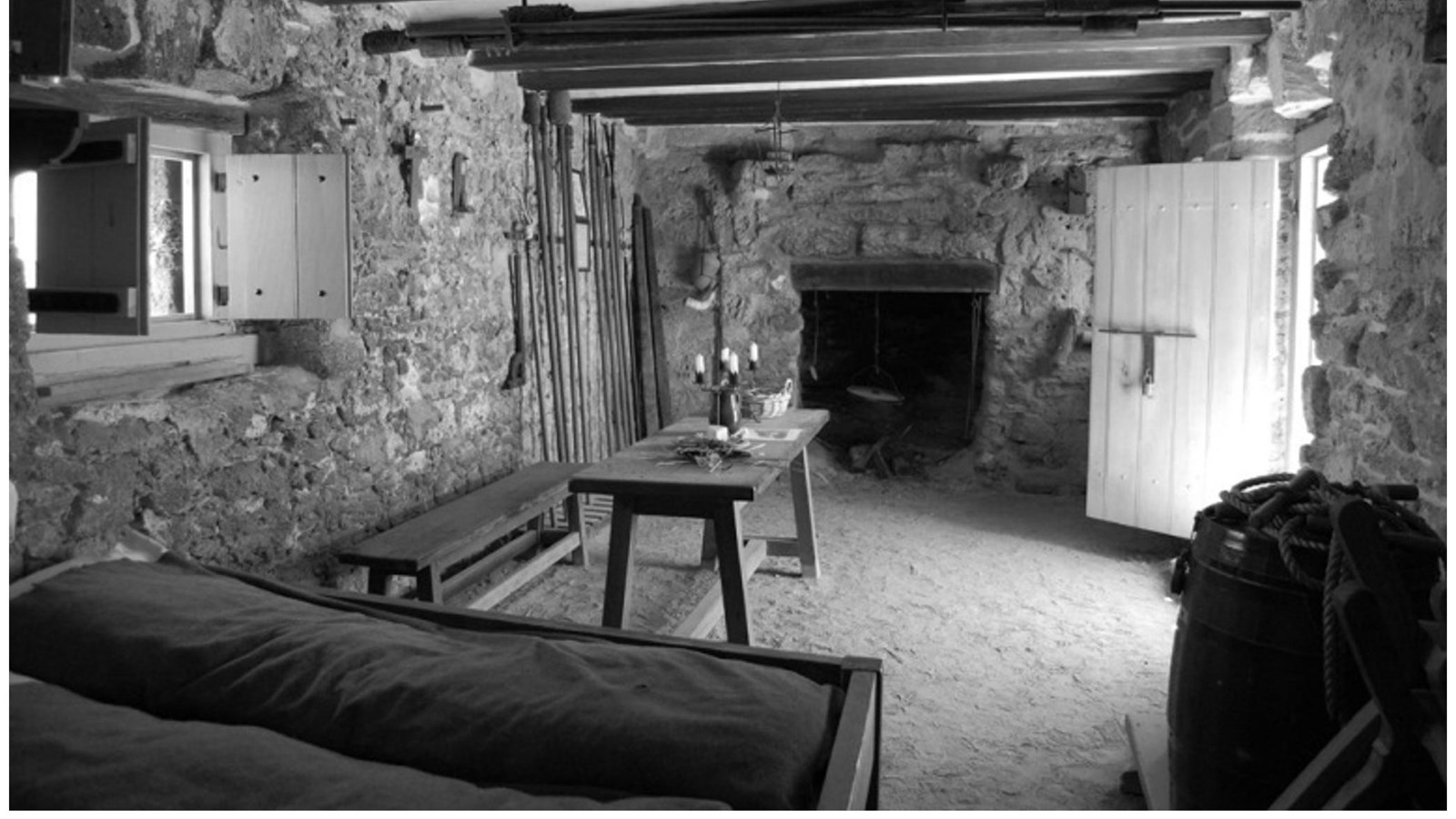 Black & white photo showing beds, fireplace, door, window, table and bench.