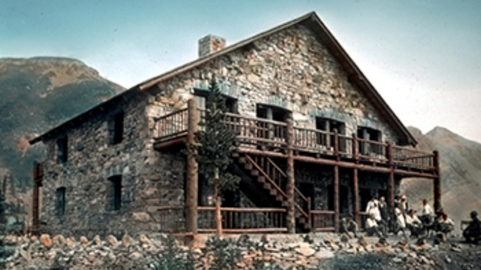 A stone two-story building with a wooden balcony and downstairs porch sit on a rocky hill.