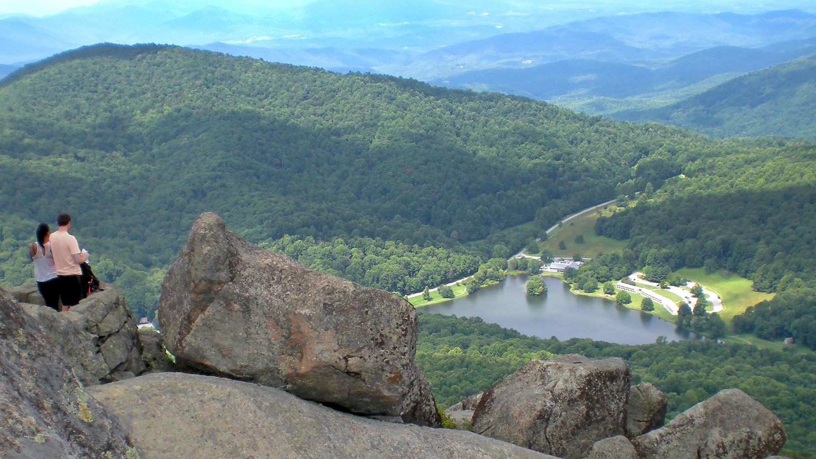Two hikes stand among large boulders on a high peak, with a small lake and logde in the valley below