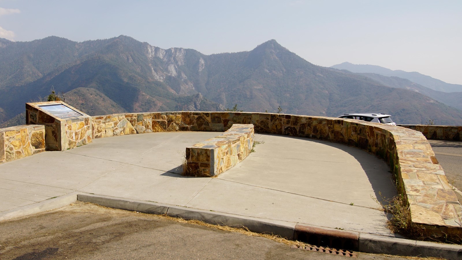 A curving rock wall sits above an asphalt highway. In the distance tall crags of rock can be seen