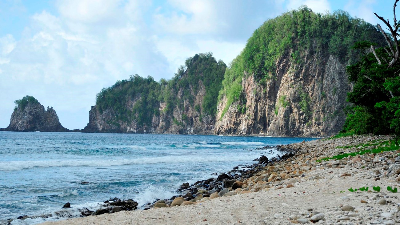 Outdoors; landscape view of sandy beach foreground, blue waters, and steep cliffs in background.  