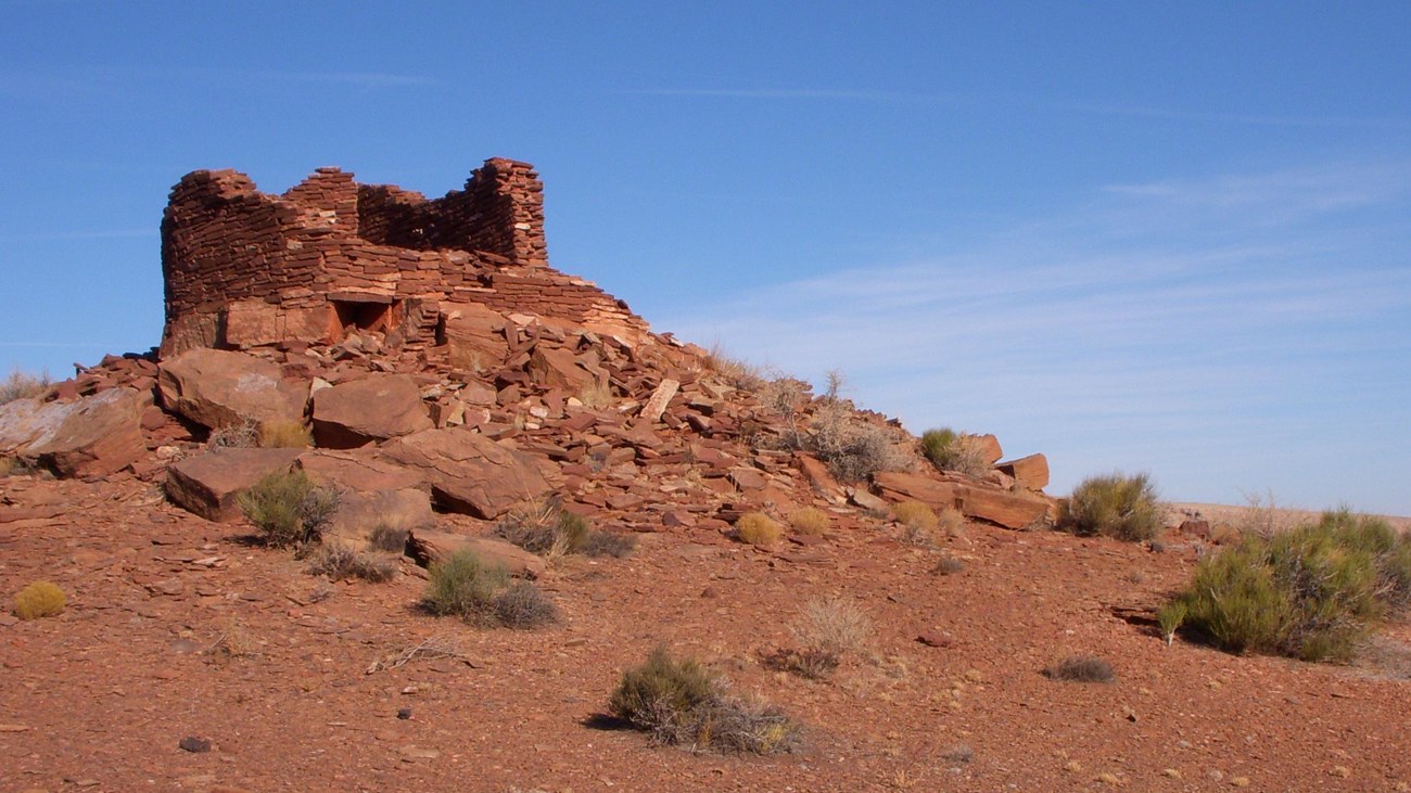 The remains of a small stone pueblo on the top of a small red sandstone hill.