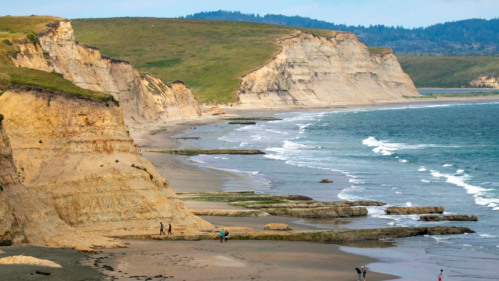 Beachgoers enjoy a sandy beach backed by sandstone cliffs as small waves wash in from the right..