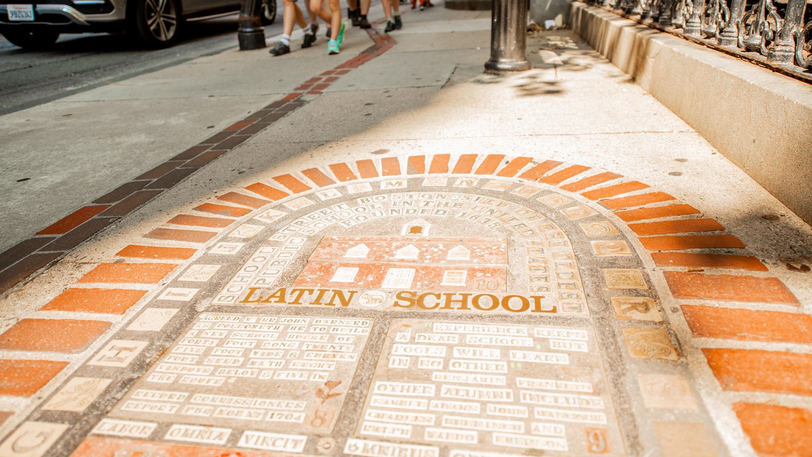 Photograph of a mosaic set in a sidewalk depicting a schoolhouse and other education elements.