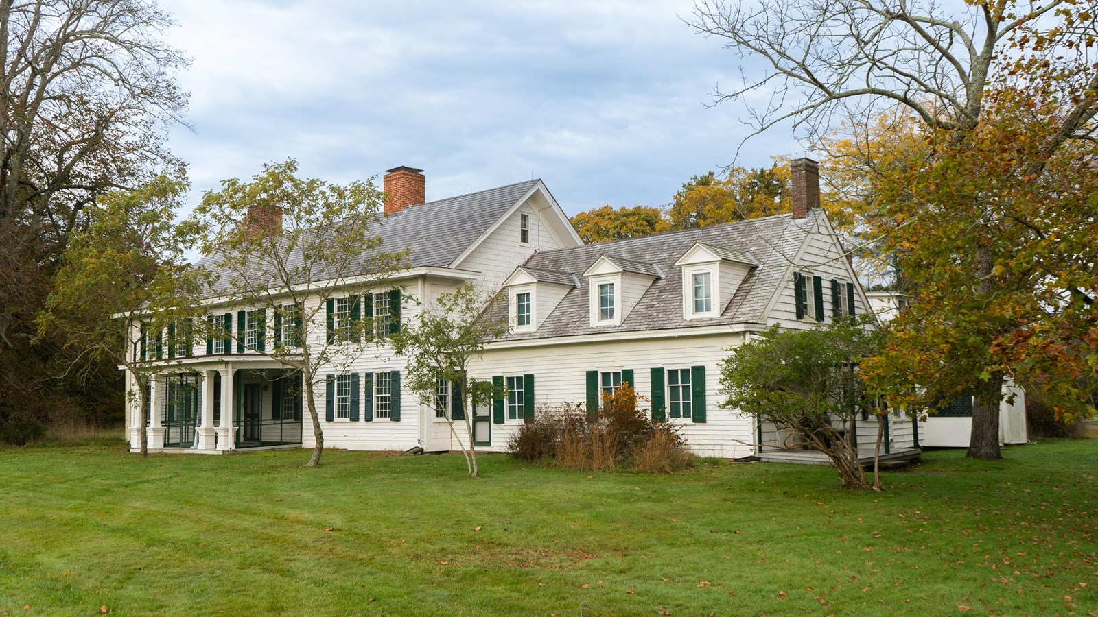 A white colonial estate with green shutters is surrounded by trees with green grass lawn.