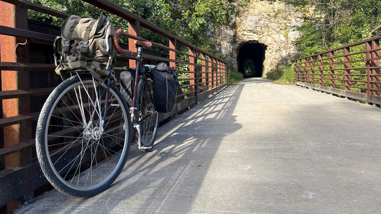 A bike strapped with bags leans on bridge railings with a tunnel through a cliff in background