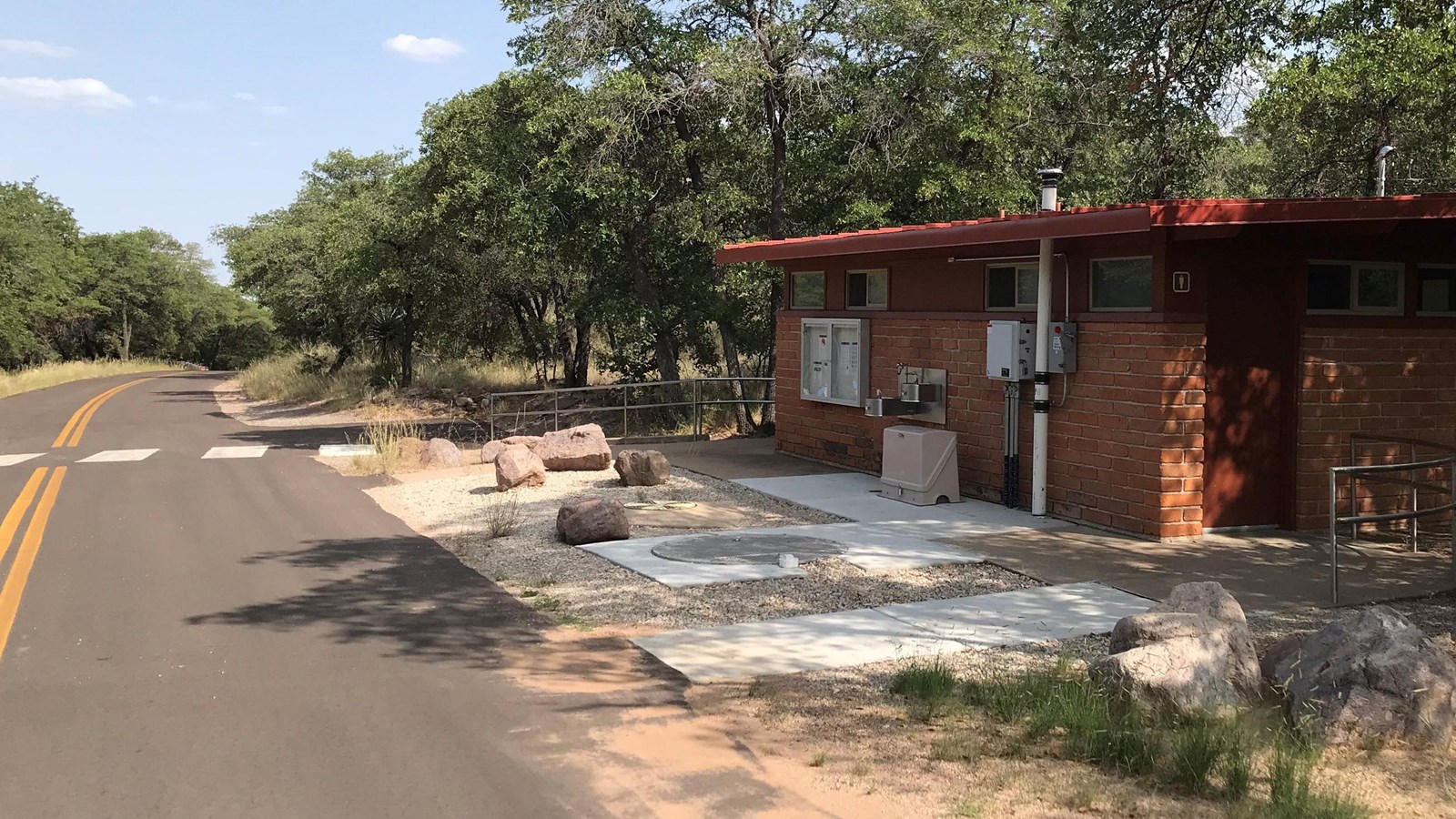 Red brick building with restrooms on either side and a single water fountain located next to road.