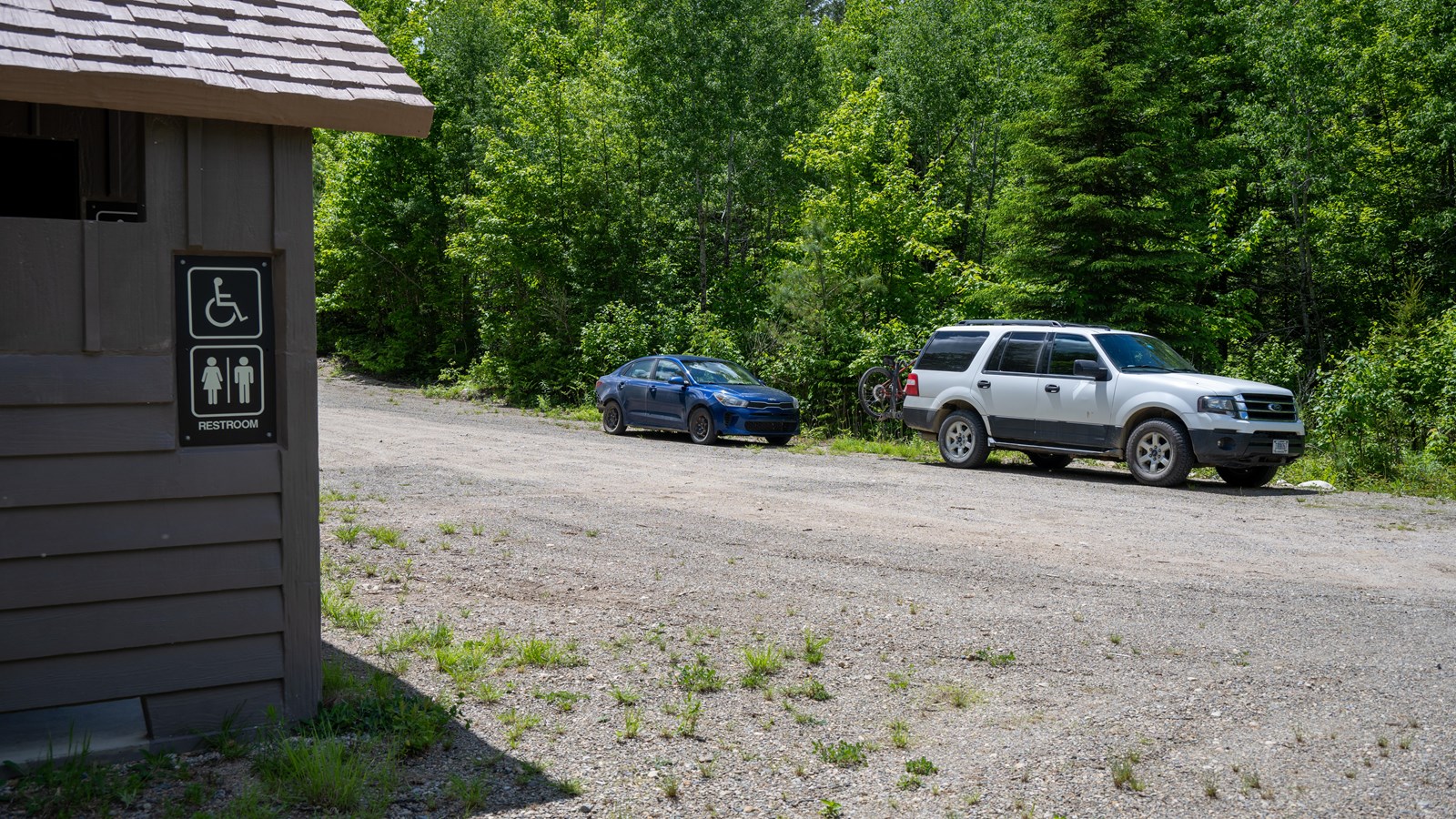 A brown vault toilet in the foreground with two cars parallel parked in a gravel park space.