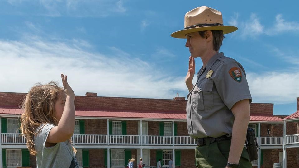 A ranger and child stand facing each other; both have their right hand in the air.