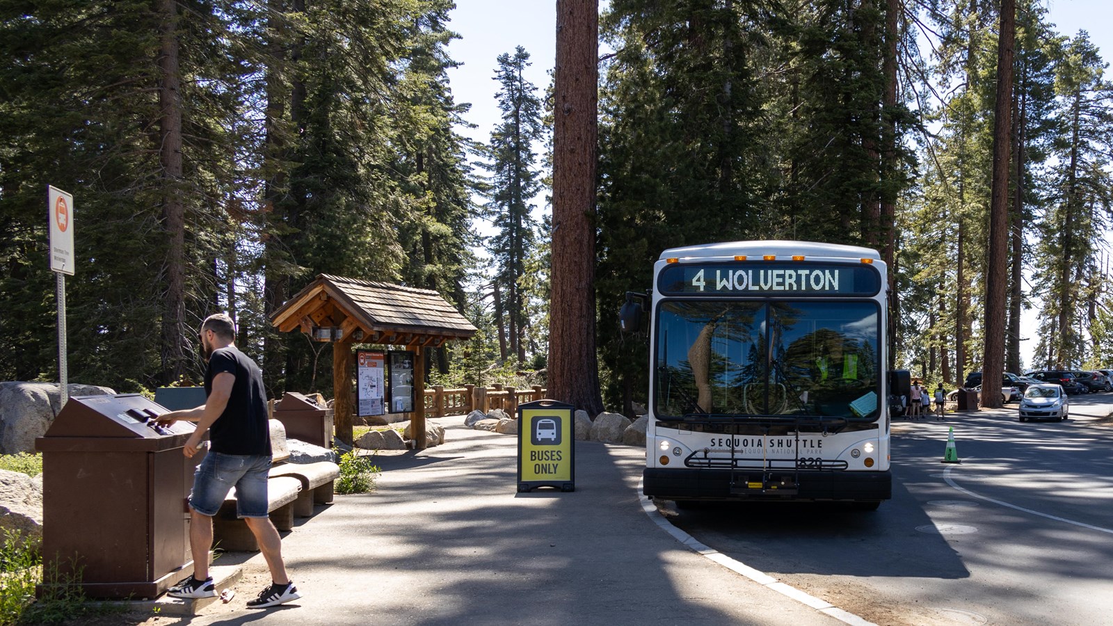 A shuttle bus arrives at a bus stop with a wooden kiosk. A man is opening a brown trashcan.