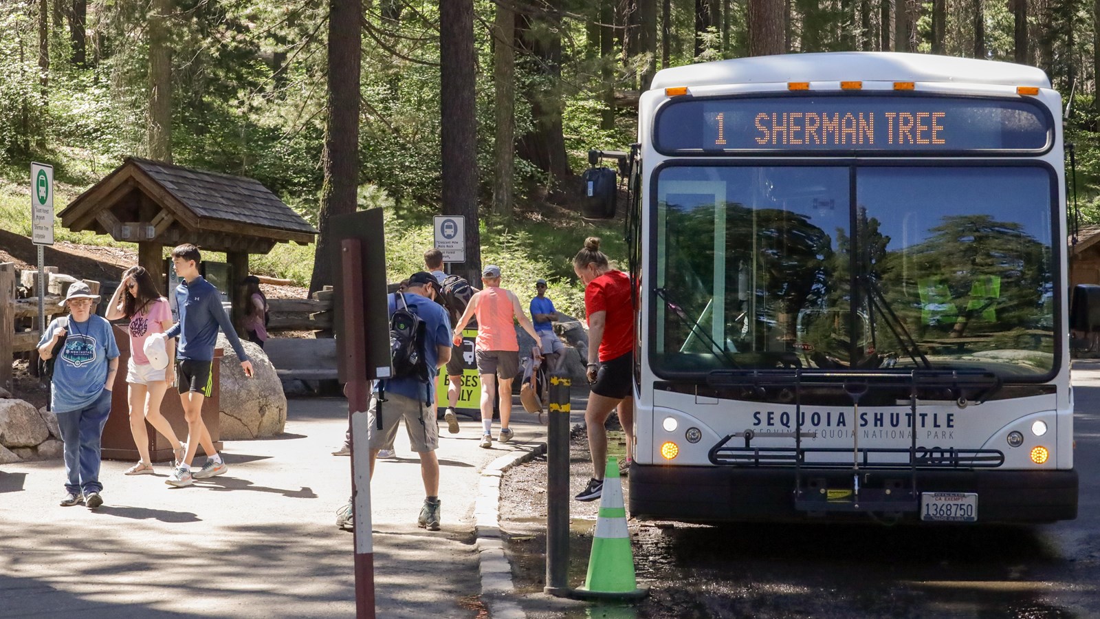 A crowd steps off a white shuttle bus. There is a shuttle stop with a wooden kiosk in the background