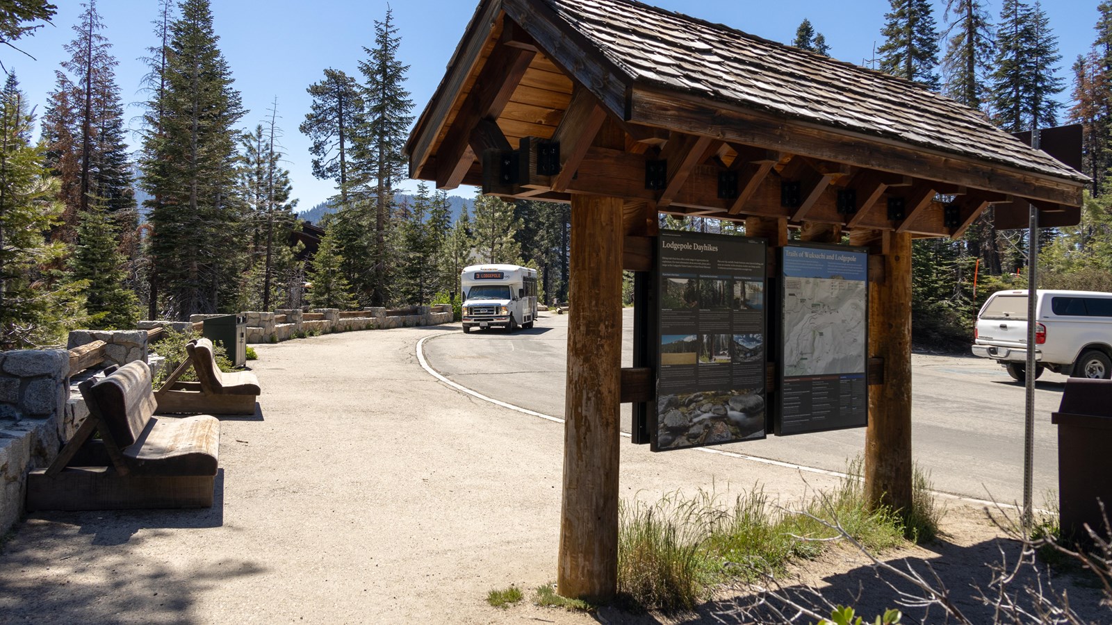 A white shuttle passenger van approaches a wooden kiosk and wooden benches.