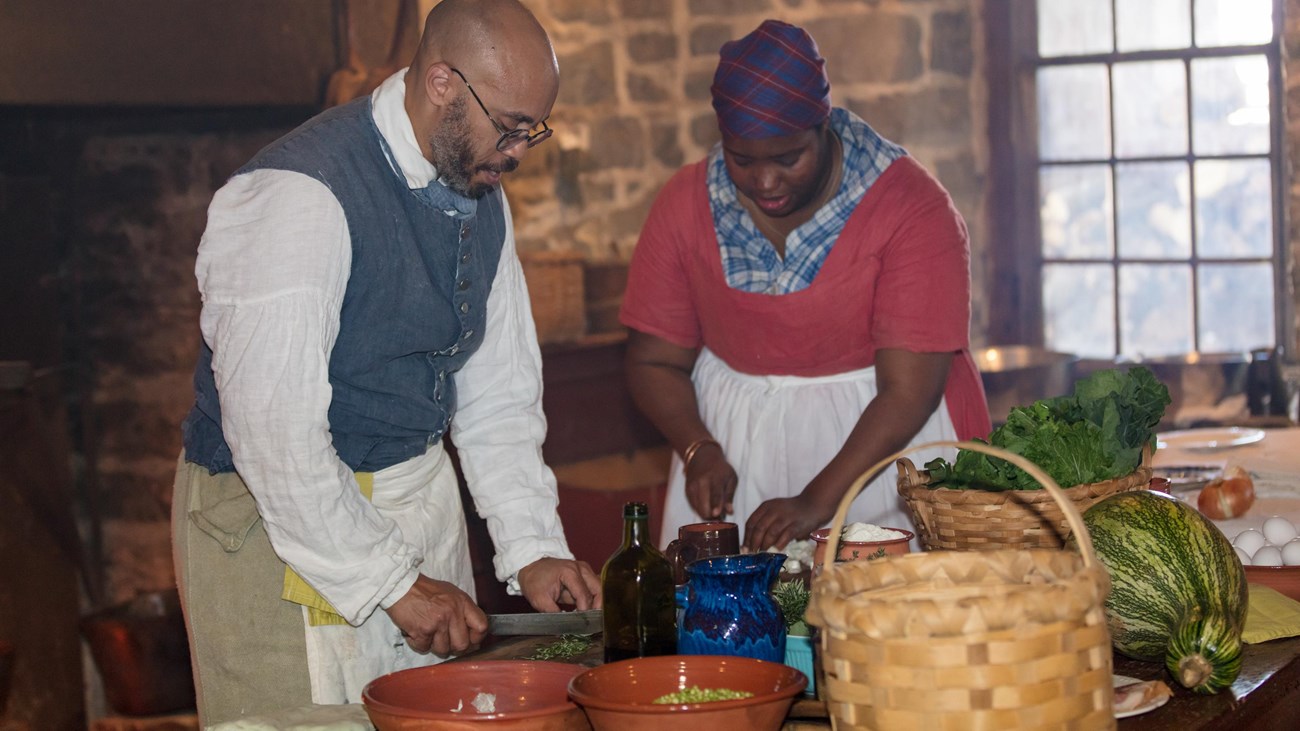 A man and woman portray enslaved cooks preparing food in a restored 1800s kitchen.