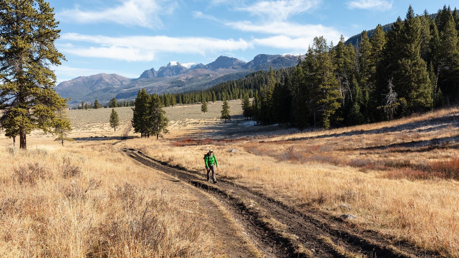 A person hikes along a muddy trail surrounded by dry grasses with mountains in the distance.