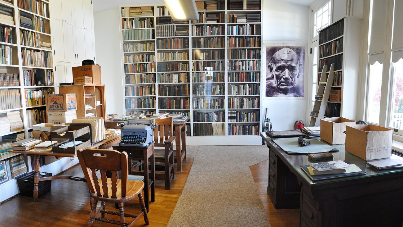 view of a study with several desks, a chair, and three walls with floor to ceiling bookshelves.