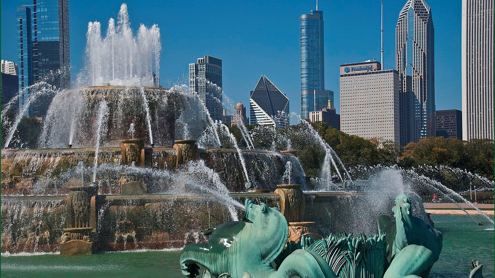 A large water fountain with a green sculpture in front and tall buildings and blue sky behind.