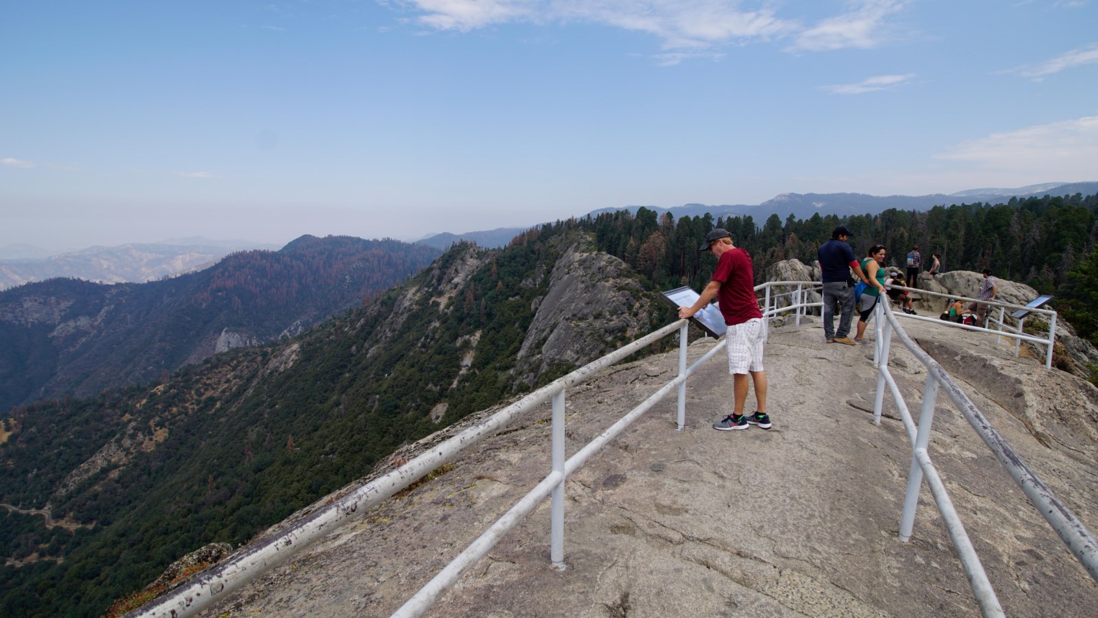 Protective rails sit atop a granite rock. In the distance are many mountains 