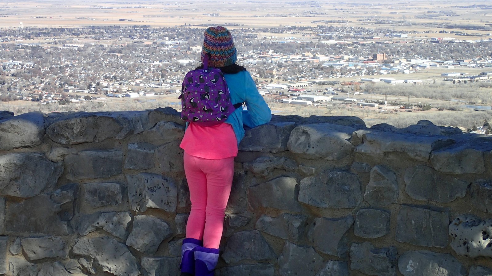 A girl stands at a stone wall overlooking a distant valley filled with homes and businesses.