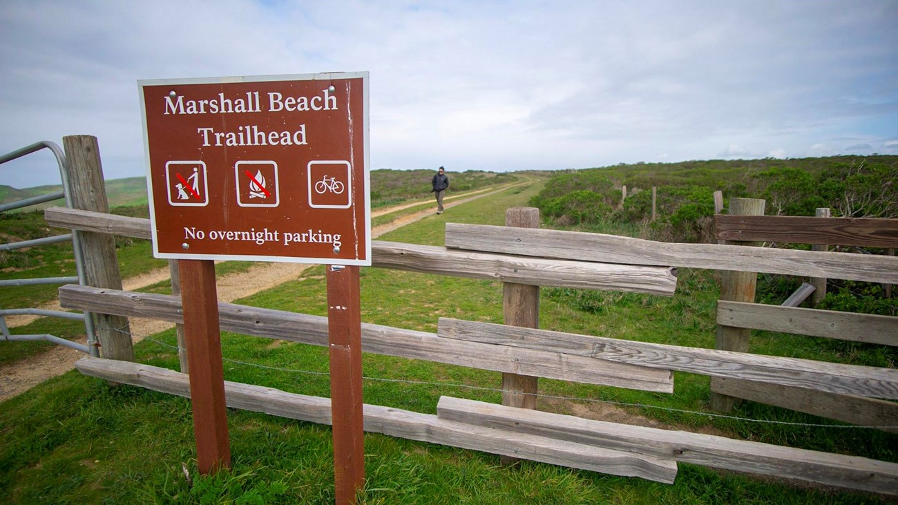 A brown trailhead sign alongside a split rail fence with a hiker on a broad trail in the background.