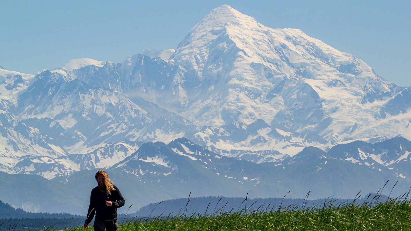 A huge snow covered mountain rises high above smaller peaks, while a person walks toward the camera