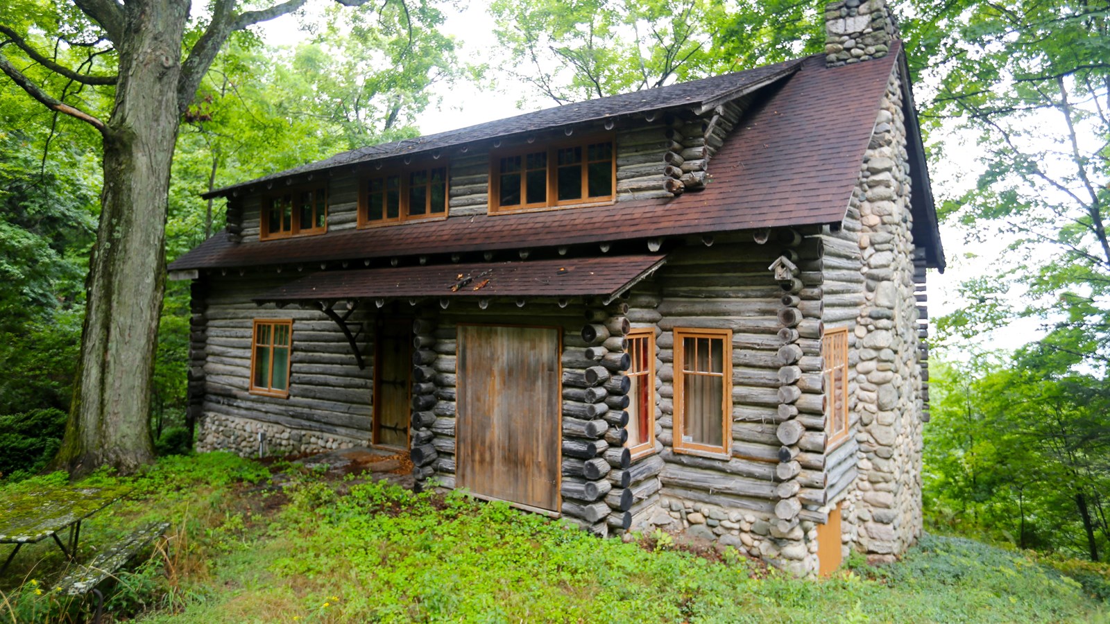 Hand hewn log cabin with three low dormer windows and covered porch surrounded by trees
