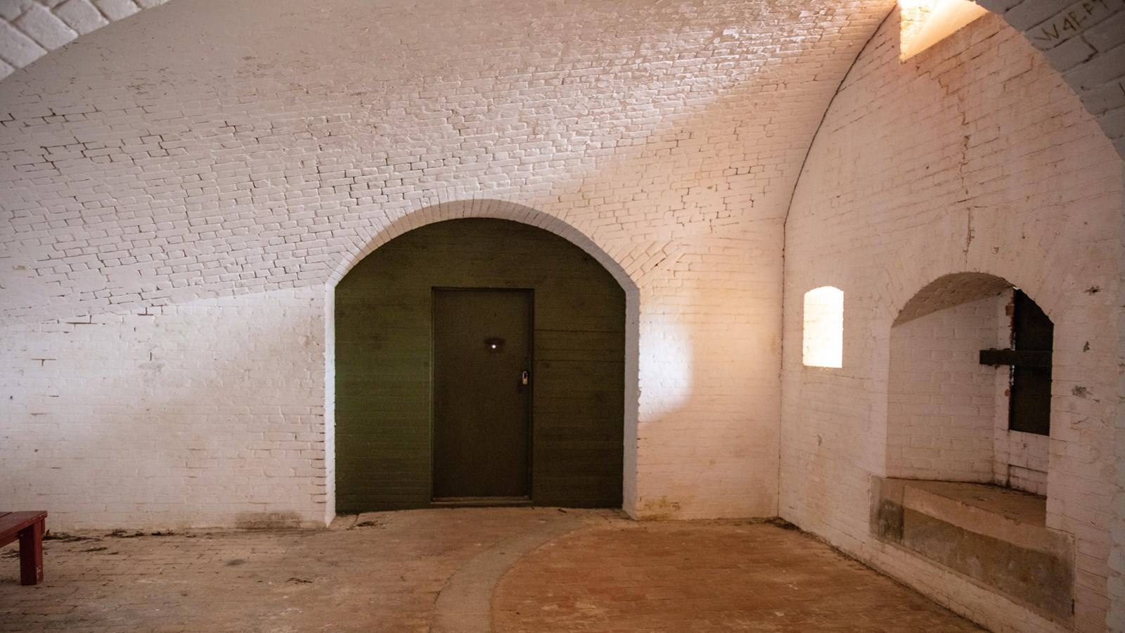 A large arched wooden door stands in a brick wall adjacent to a wall with a hole for cannon.