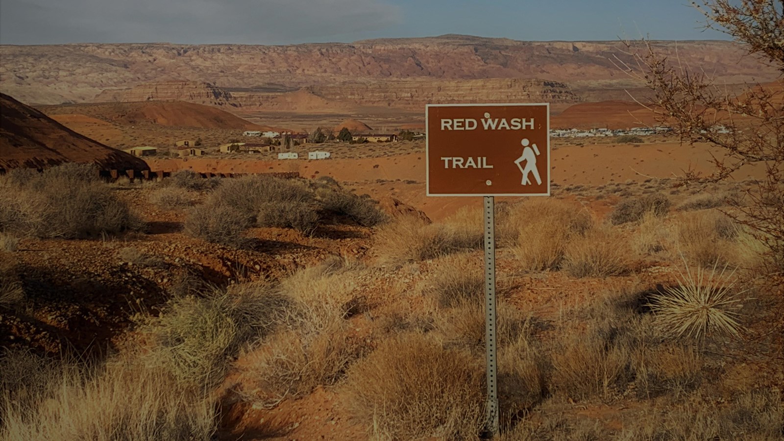 Brown sign with hiking symbol on it next to dip in desert landscape.