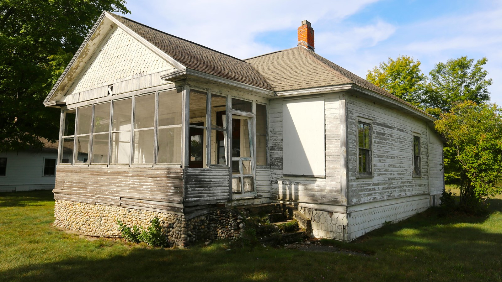 Graying paint covers a single-story cottage with river rock foundation supporting an enclosed porch