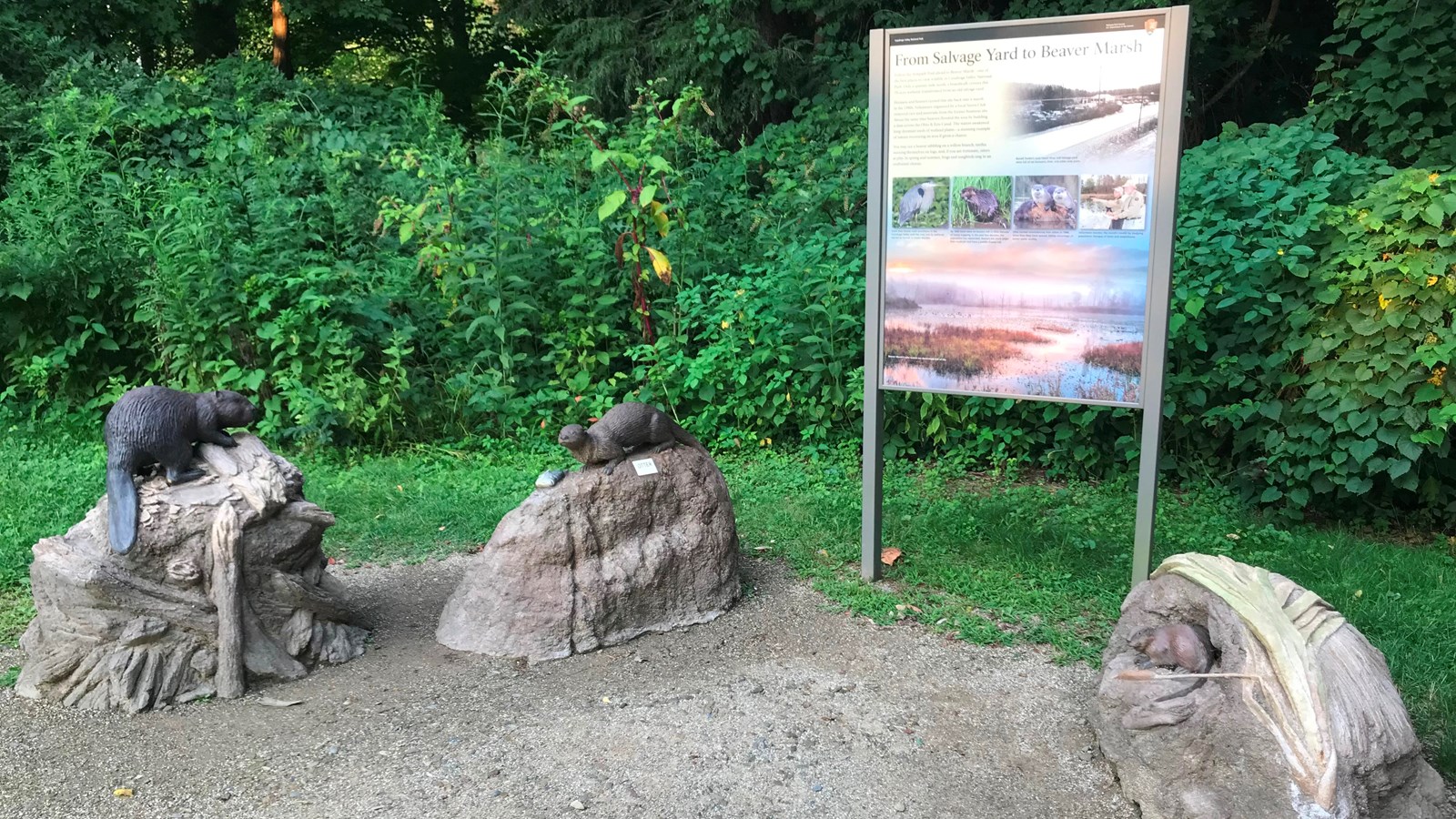 Three sculptures sit on raised mounds around an upright graphic panel with bushes in the background.