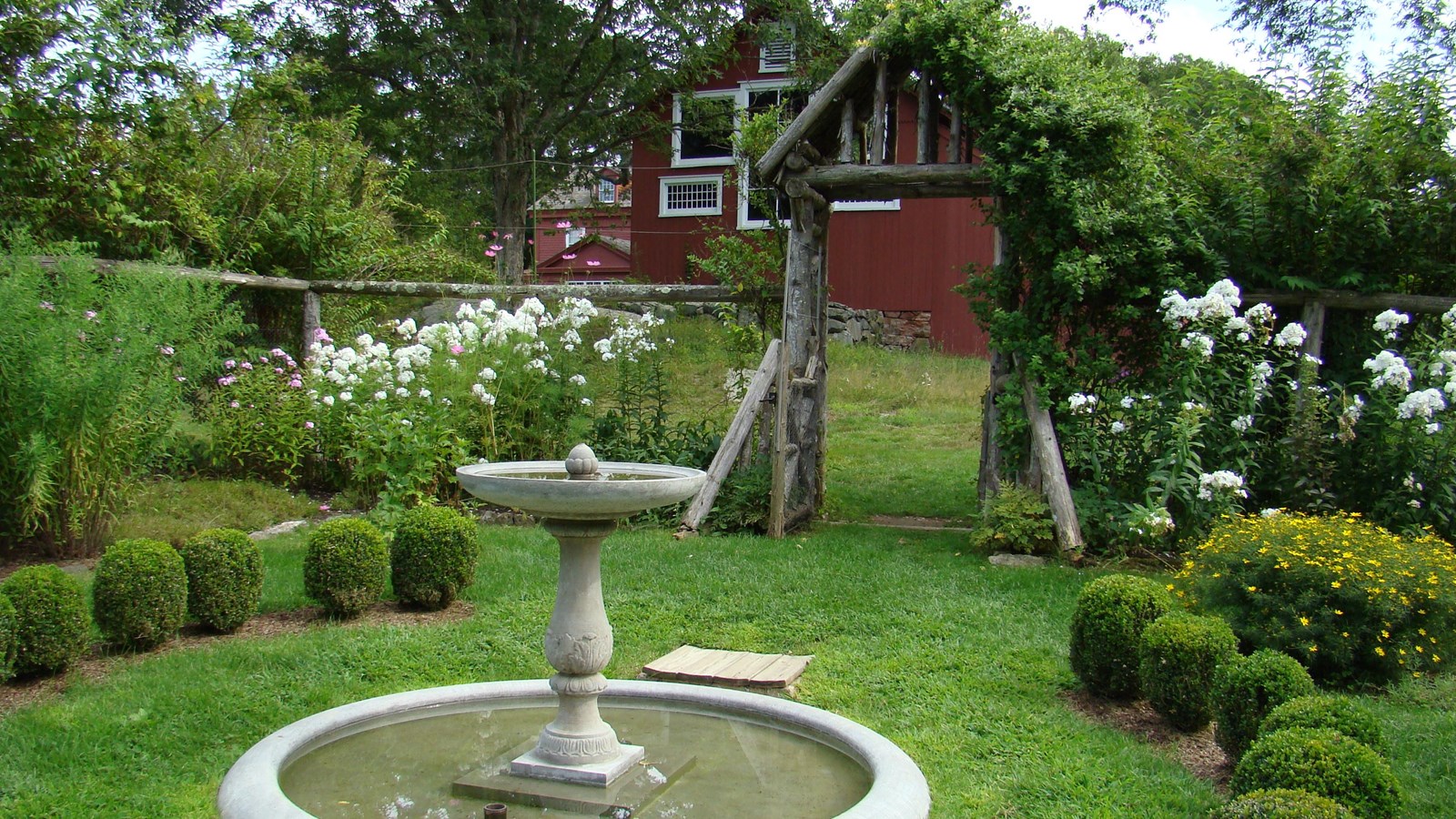 A water fountain in the middle surrounded by small green bushes and a wooden gate in the background.