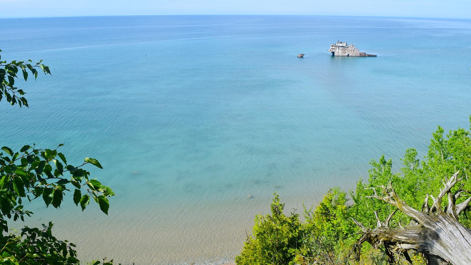 A gray metal hulk sticks out of the turquoise water with green trees in the foreground