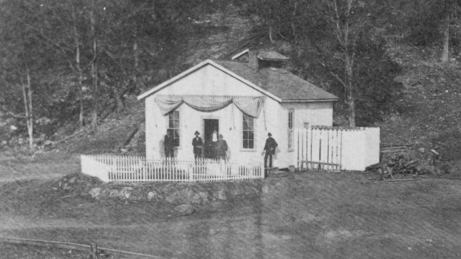White building in a grassy area with surrounded by fences. Five men stand around the doorway.