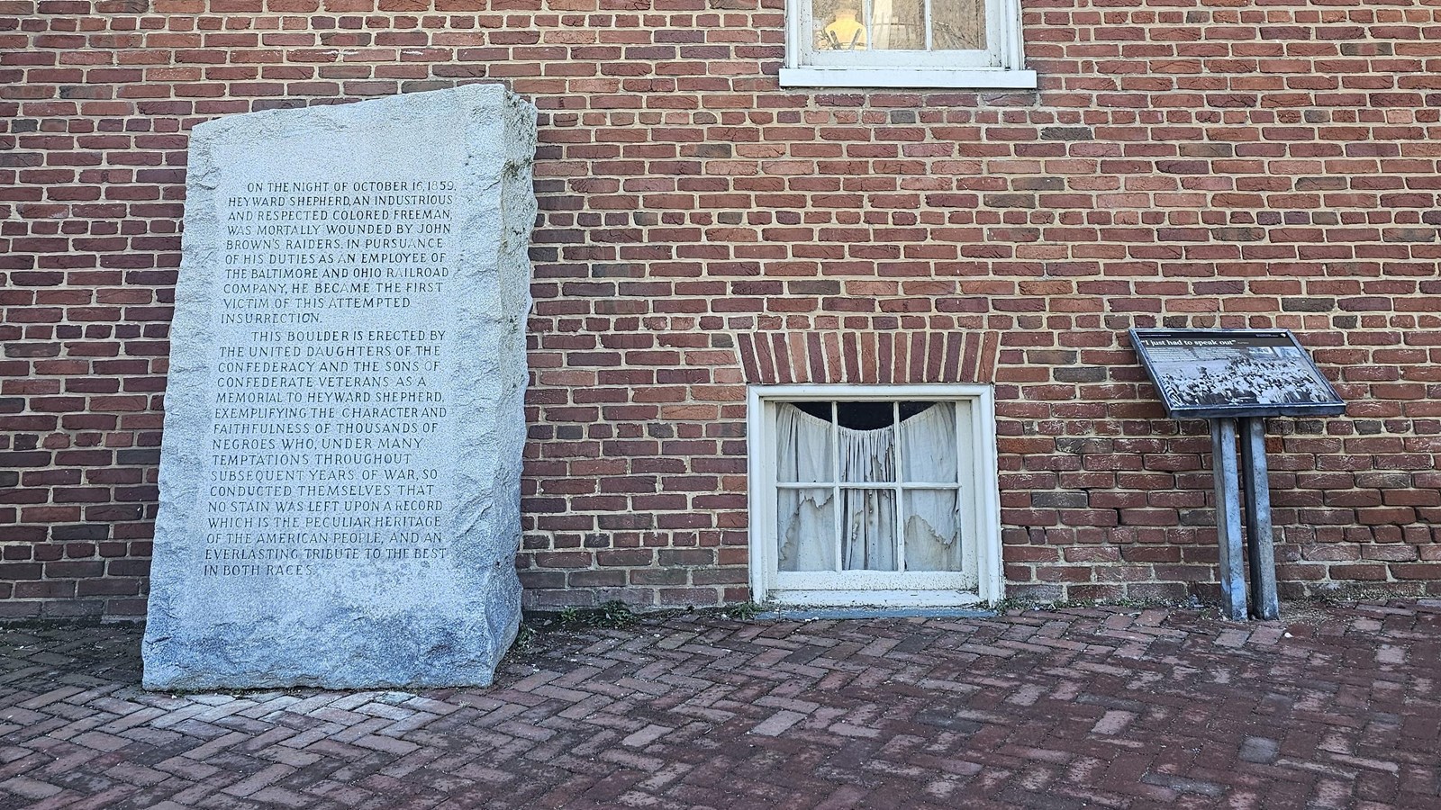 A stone monument and sign stand side by side in front of a brick building.