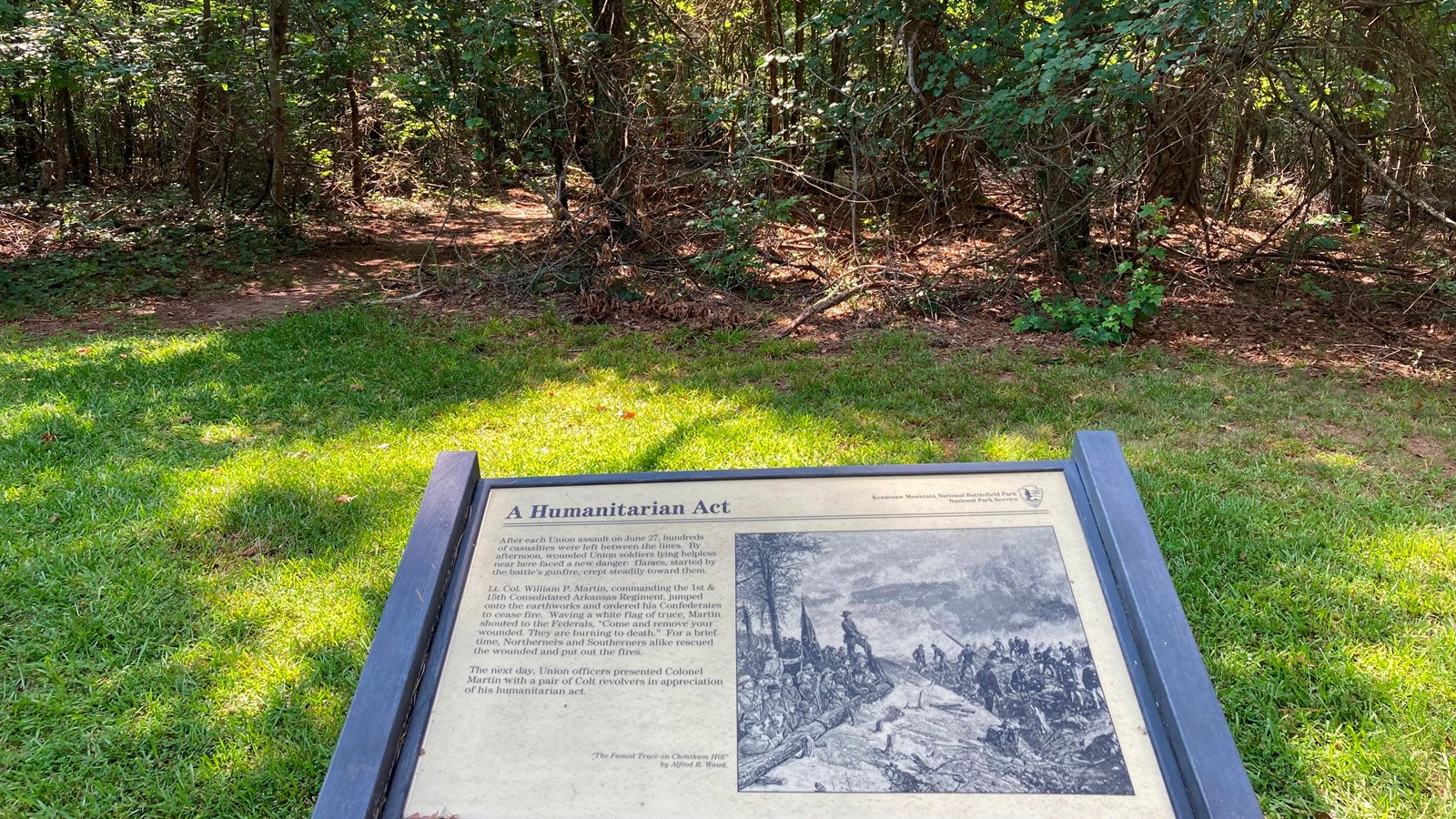 A grassy section and line of trees. In the foreground is a sign titled \'An Act of Humanity\'