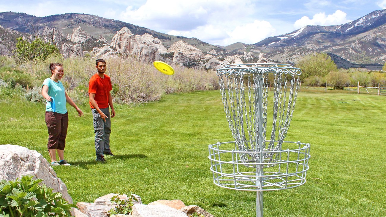 Two people watch a yellow disc flying toward the metal disc golf basket, castle rocks in the back.